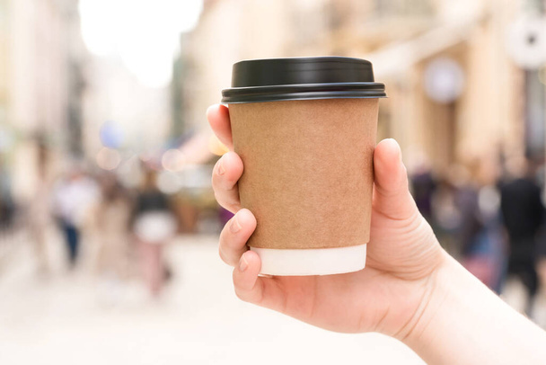 Young woman's hand holding kraft paper coffee cup against blurred city street as background. Coffee take away concept. Kraft paper coffee cup with plastic lid. Mockup - Foto, afbeelding