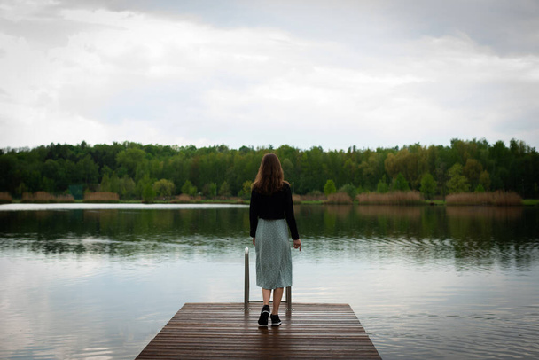 A young slender woman in cloudy weather walks to the edge of a wooden pier - Foto, Bild