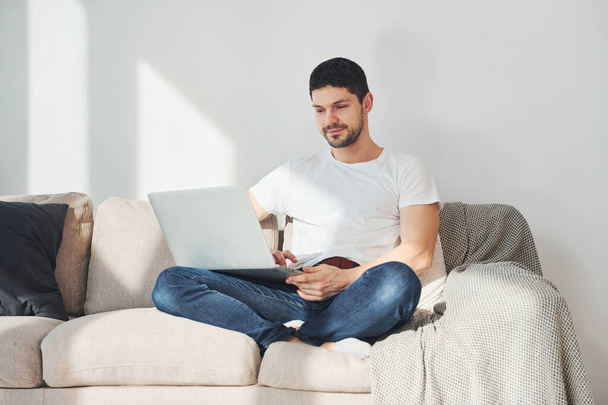 Man in white shirt and jeans sitting on bed with laptop indoors. - 写真・画像