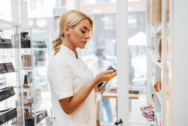 Young and attractive female pharmacist working in a drugstore. She is confident and serious. - Фото, изображение