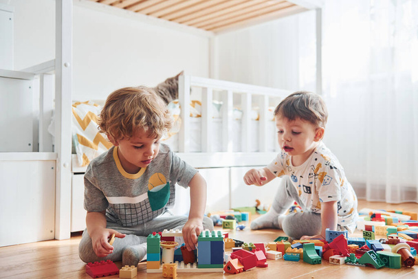 Two little boys have fun indoors in the bedroom with plastic construction set. Cat sits behind them. - Foto, Imagem