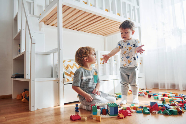 Two little boys have fun indoors in the bedroom with plastic construction set. - 写真・画像