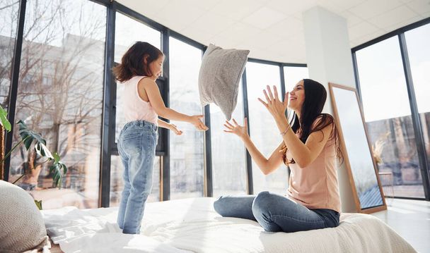 Having fun with pillow on bed. Young mother with her daughter spending weekend together in room. - Photo, Image