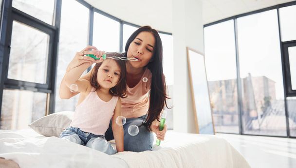 Young mother with her daughter blowing bubbles together in bedroom. - 写真・画像