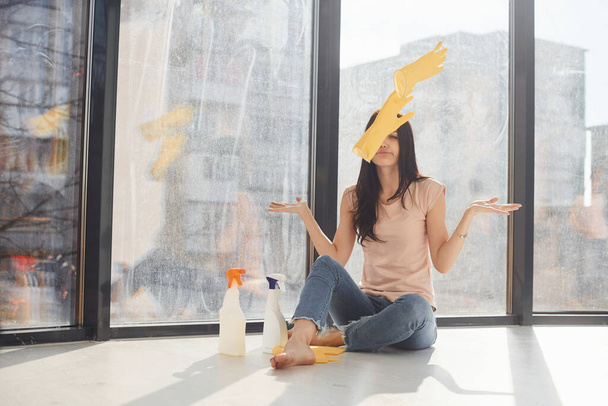 Yorgun bir kadın pencere eşiğinde oturur. Elinde temizleme spreyi şişeleriyle havaya eldiven fırlatır.. - Fotoğraf, Görsel