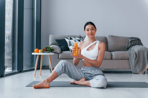 Holds glass with orange juice in hands. Young woman with slim body shape in sportswear have fitness day indoors at home. - Photo, Image