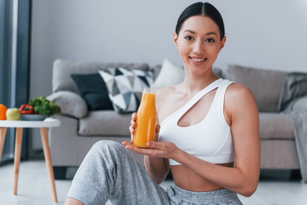 Holds glass with orange juice in hands. Young woman with slim body shape in sportswear have fitness day indoors at home. - Фото, зображення
