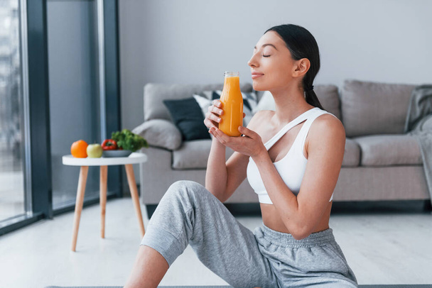 Holds glass with orange juice in hands. Young woman with slim body shape in sportswear have fitness day indoors at home. - Foto, imagen