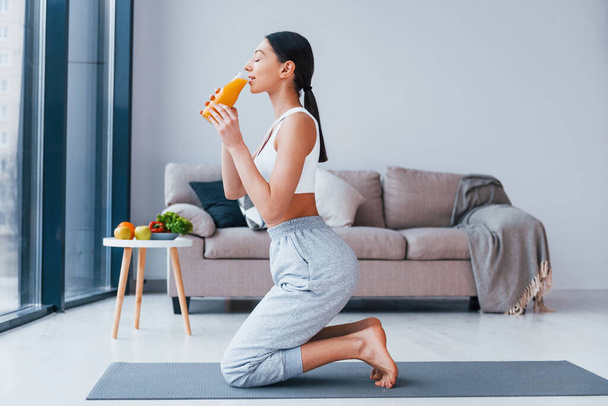 Holds glass with orange juice in hands. Young woman with slim body shape in sportswear have fitness day indoors at home. - Foto, Imagem
