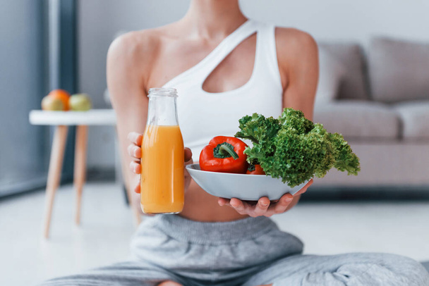 With healthy food and orange juice in hands. Young woman with slim body shape in sportswear have fitness day indoors at home. - Фото, зображення