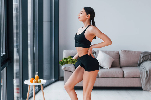 holds healthy food in hands. Young woman with slim body shape in black sportswear have fitness day indoors at home. - Foto, Imagen