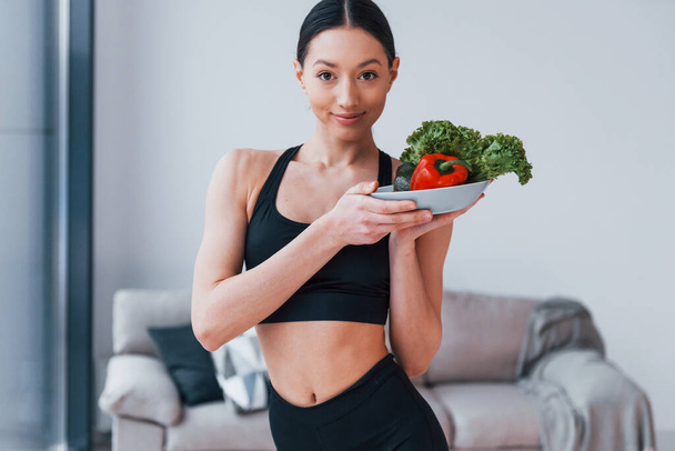 holds healthy food in hands. Young woman with slim body shape in black sportswear have fitness day indoors at home. - Fotografie, Obrázek