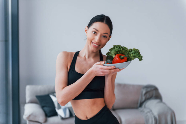 holds healthy food in hands. Young woman with slim body shape in black sportswear have fitness day indoors at home. - Фото, изображение