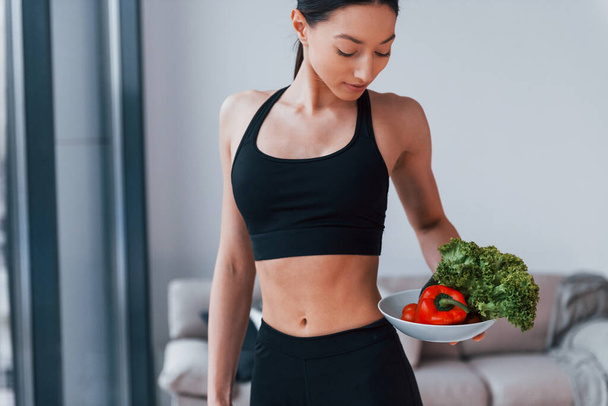 holds healthy food in hands. Young woman with slim body shape in black sportswear have fitness day indoors at home. - Фото, изображение