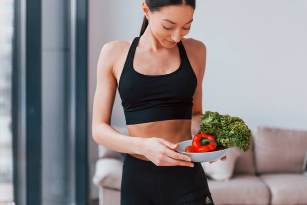 holds healthy food in hands. Young woman with slim body shape in black sportswear have fitness day indoors at home. - Fotó, kép