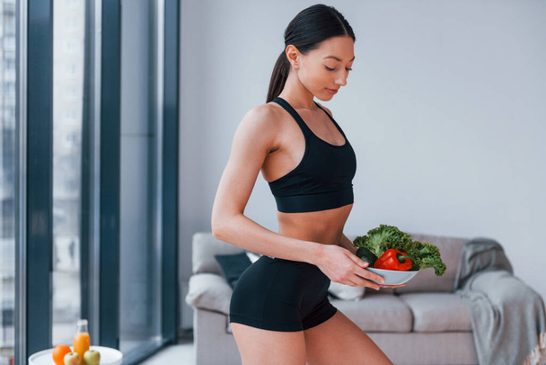 holds healthy food in hands. Young woman with slim body shape in black sportswear have fitness day indoors at home. - Zdjęcie, obraz