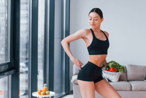 holds healthy food in hands. Young woman with slim body shape in black sportswear have fitness day indoors at home. - Photo, image