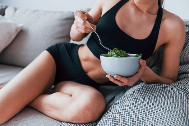 Young woman with slim body shape in sportswear sits on sofa and eats healthy diet food indoors at home. - Valokuva, kuva