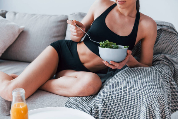 Young woman with slim body shape in sportswear sits on sofa and eats healthy diet food indoors at home. - Fotografie, Obrázek