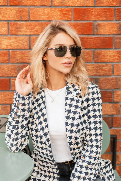 Pretty glamour young woman with sunglasses in fashion outfit sits on the street near red brick wall - 写真・画像