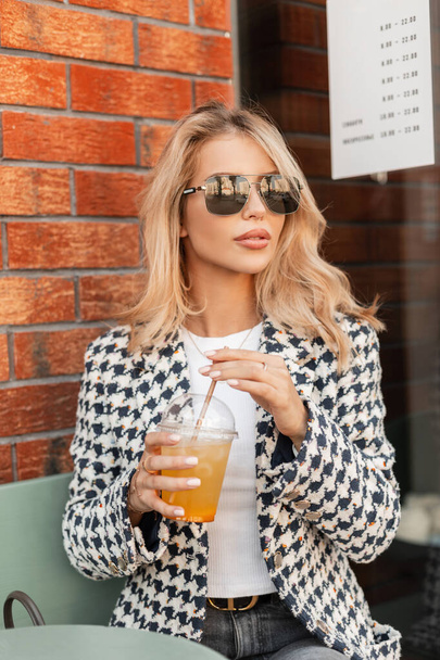 Stylish pretty blonde business lady with sunglasses in fashionable outerwear sits in a summer cafe and drinks a cocktail juice - Photo, Image