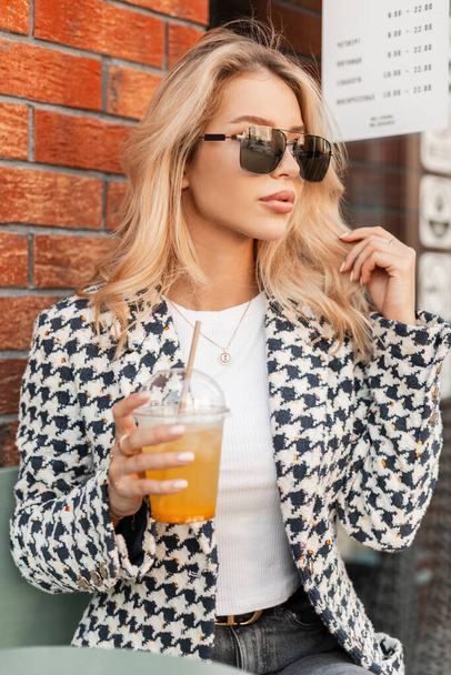 Beautiful sexy blonde woman with cool sunglasses in a fashionable jacket coat sits on a summer terrace near a red brick wall and drinks lemonade - Zdjęcie, obraz