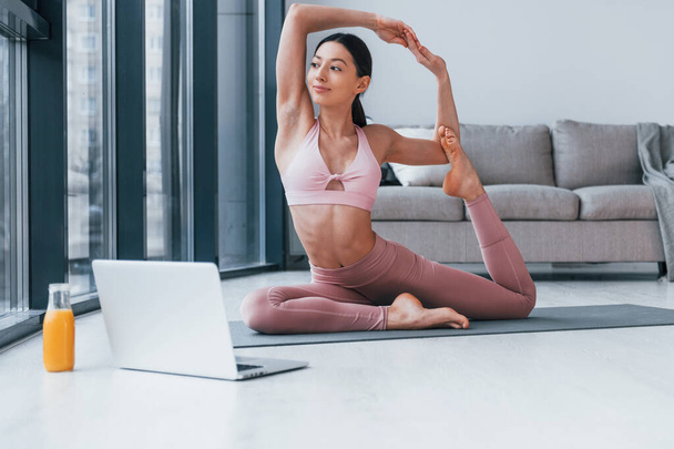 Does exercises on mat on the floor with laptop and glass of orange juice on it. Young woman with slim body shape in sportswear have fitness day indoors at home. - Photo, Image