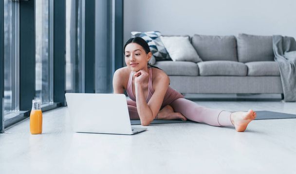 Young woman with slim body shape in sportswear sits on mat with orange juice and laptop indoors at home. - Фото, изображение