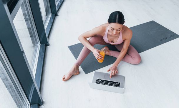 Young woman with slim body shape in sportswear sits on mat with orange juice and laptop indoors at home. - Fotografie, Obrázek