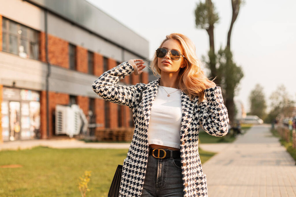 Beautiful glamorous young blonde woman in fashionable outerwear with sunglasses and a handbag walks in the city at sunset. Summer women's clothing urban style look - Fotografie, Obrázek
