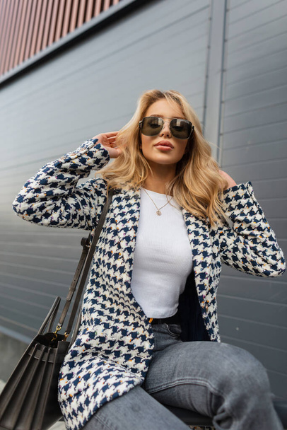 Fashion blonde woman hipster with vintage sunglasses in stylish elegance clothes with jacket, jeans and handbag sits on the street. Urban female style outwear look - 写真・画像