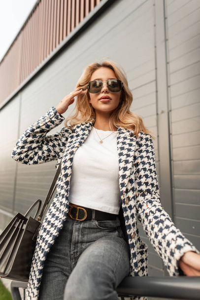 Stylish beautiful young blonde girl with fashionable sunglasses in trendy elegant clothes with a jacket, a T-shirt, jeans and a handbag sits in the city near a modern wall - Fotó, kép