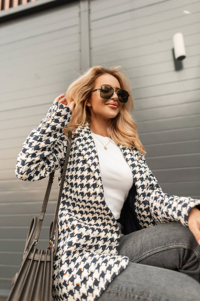 Chica joven glamorosa feliz en ropa elegante de moda con una chaqueta, denim, un bolso y gafas de sol se sienta en la ciudad cerca de la pared moderna - Foto, Imagen