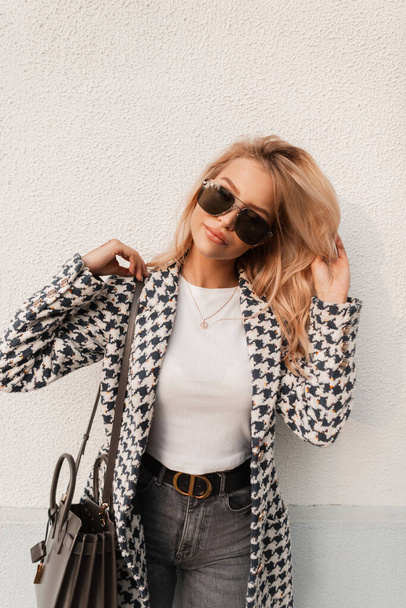 Stylish beautiful young woman in fashionable clothes with a jacket, sunglasses and a handbag stands near a white wall in the city. Women's elegant streetwear style - 写真・画像