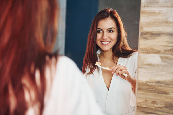 Beautiful young woman standing in bathroom near the mirror and brushing her teeth. - Foto, Imagen