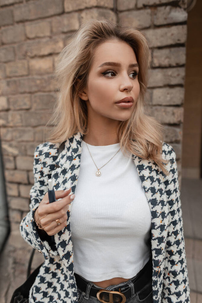 Beautiful young blonde girl in fashionable clothes in the city near a brick vintage wall - 写真・画像