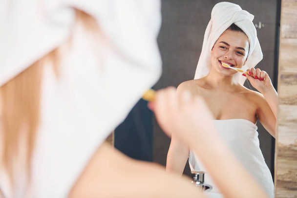 Beautiful young woman standing in bathroom near the mirror and brushing her teeth. - 写真・画像