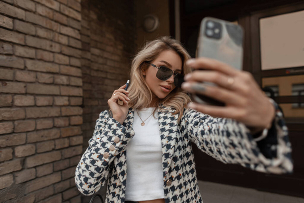 Stylish young woman in fashionable clothes with sunglasses and an e-cigarette walks in the city and takes a selfie photo on phone - Foto, afbeelding
