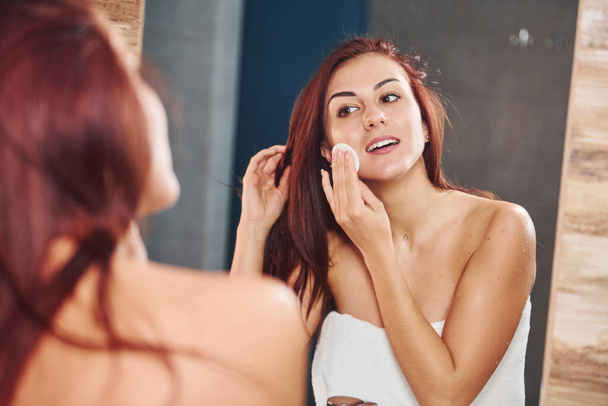 Beautiful young woman standing in bathroom, looking into the mirror and taking care of her face. - Foto, Imagem