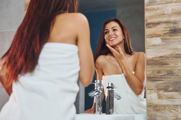 Beautiful young woman standing in bathroom, looking into the mirror and taking care of her face. - Foto, imagen