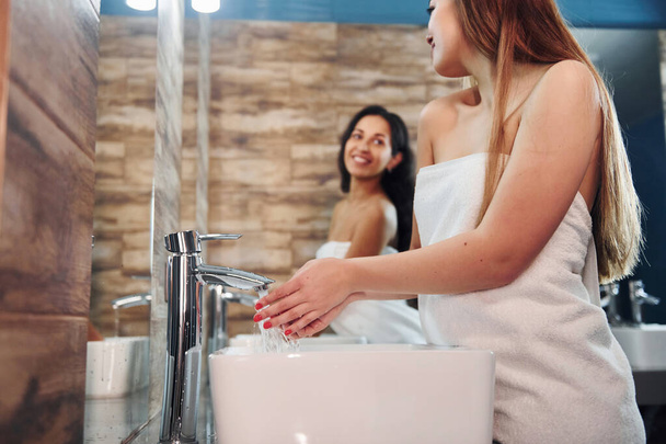Krásná mladá žena stojí v koupelně u zrcadla a myje si ruce. Přátelé za. - Fotografie, Obrázek
