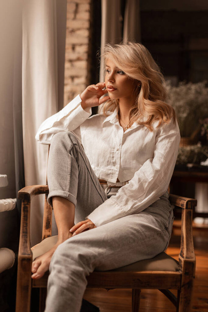 Pretty young sexy woman with blond hair in fashionable clothes with a white linen shirt and jeans sits in a wooden vintage armchair - Fotografie, Obrázek