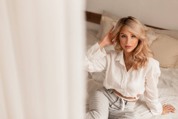 Elegant beautiful blonde woman with blond hairstyle in a fashionable white shirt and vintage jeans sits and rests on the bed in the bedroom - Photo, image