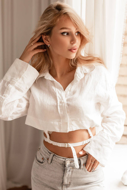 Beautiful girl with blond hair in elegant fashion clothes with a white shirt and high waist jeans indoors at home - 写真・画像