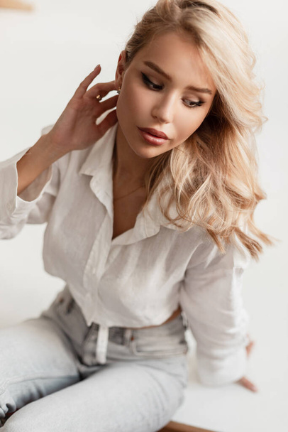Beautiful young blonde girl in a fashionable white top with jeans sitting on a white background indoors - 写真・画像