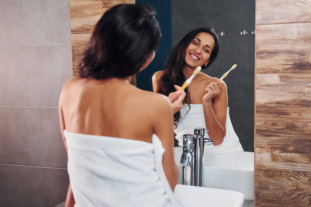 Beautiful young brunette standing in bathroom near the mirror and brushing her teeth. - Valokuva, kuva