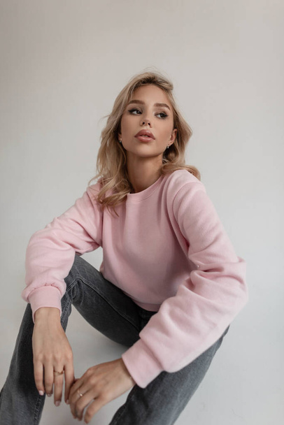 Beautiful young woman hipster with blond hair in a fashion pink sweatshirt and vintage gray jeans is sitting on the floor in the studio - Photo, Image