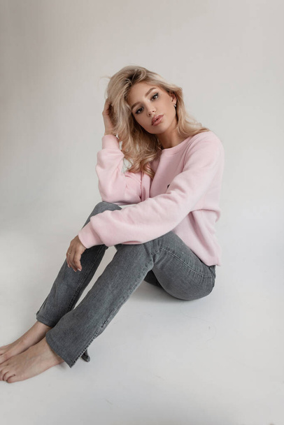 Glamorous beautiful slender girl with blond hair in a fashionable pink sweatshirt and vintage stylish jeans sits on a white background in the studio - Фото, зображення