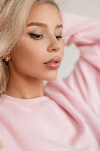 Female stylish portrait of a pretty young blonde girl model with beautiful lips and natural beauty in a fashion pink sweatshirt indoors - Zdjęcie, obraz