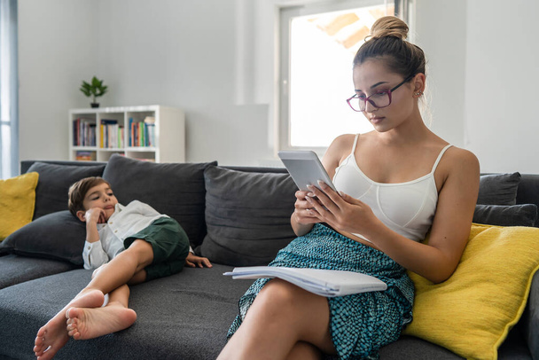 Beyaz bir kadın, elinde dijital tablet ve defter ile evdeki kanepede oturan çocuğu ya da kardeşinin yanında oturuyor. Finans okuyor ya da yaz günü evde yatırım planları yapıyor. - Fotoğraf, Görsel